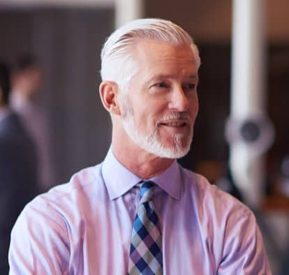 Man with gray hair and beard, wearing a light purple shirt and patterned tie, standing indoors.