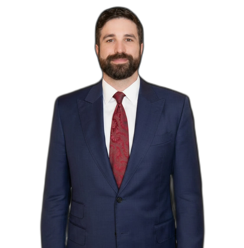 Man with dark hair and beard wearing a navy blue suit, white shirt, and red patterned tie, standing and facing forward against a plain white background.