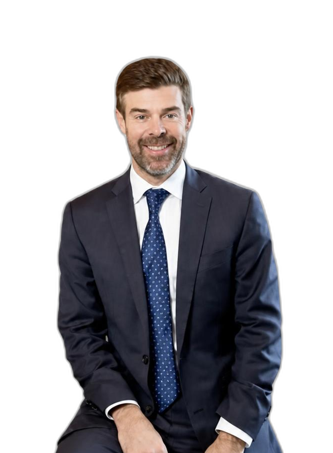 A man in a navy suit and blue polka-dot tie sits on a stool against a plain white background, smiling at the camera.