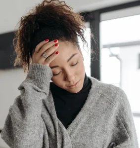 A person with curly hair and red nail polish is touching their forehead with eyes closed, appearing tired or stressed, wearing a gray sweater and black top indoors.