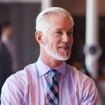 Man with gray hair and beard, wearing a light purple shirt and patterned tie, standing indoors.