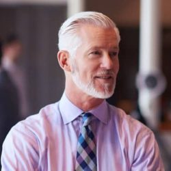 Man with gray hair and beard, wearing a light purple shirt and patterned tie, standing indoors.