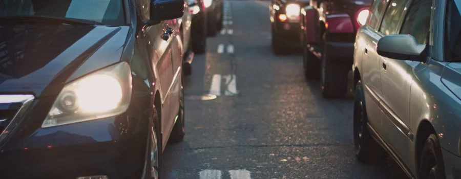 Cars are stopped in heavy traffic on a city street, with headlights on and buildings visible in the background.